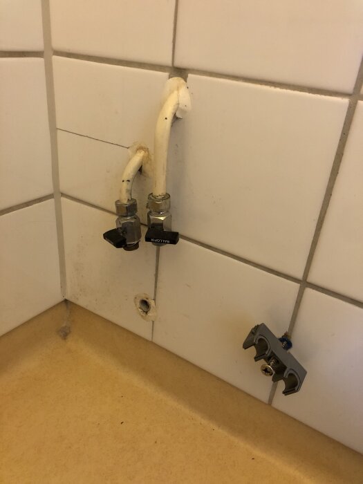 Vägg med vit kakel, två vattenrör med ventiler och ett igenpluggat avloppshål.