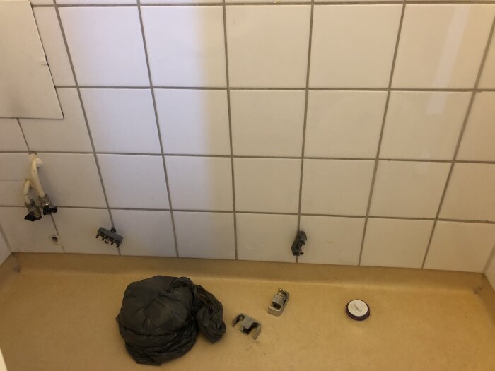 Ett gästbadrum under renovering med vita kakelplattor, avstängda vattenrör och ett igenpluggat avloppshål. Plastpåse och verktyg ligger på golvet.