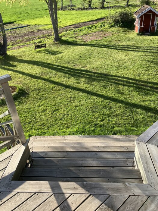Trätrappa med utsikt över grön gräsmatta, en bänk under träden och ett litet rött förråd i hörnet.
