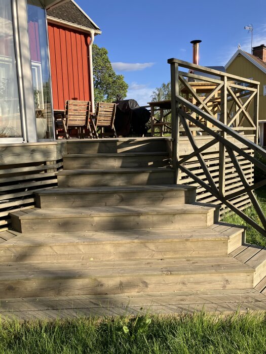 Stor altan med trätrappa och räcke, trädgårdsmöbler samt grill på en solig dag vid ett rött hus.