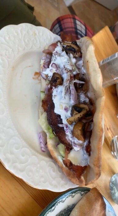 Pizzabröd fyllt med sallad, rödlök, tomat, gurka, BBQ-sås, stekt fläsk, vitlökssås och stekta champinjoner på en tallrik.