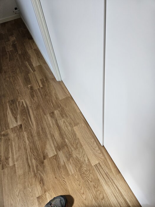 Vit parskjutdörr med en synlig glipa på cirka en centimeter, bredvid ett trägolv. En fot syns i hörnet.
