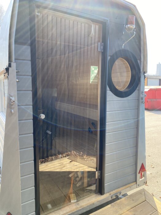 Entrésidan av en mobilbastu med öppen glasdörr, träpanel synlig inuti, och ett runt fönster på sidan.