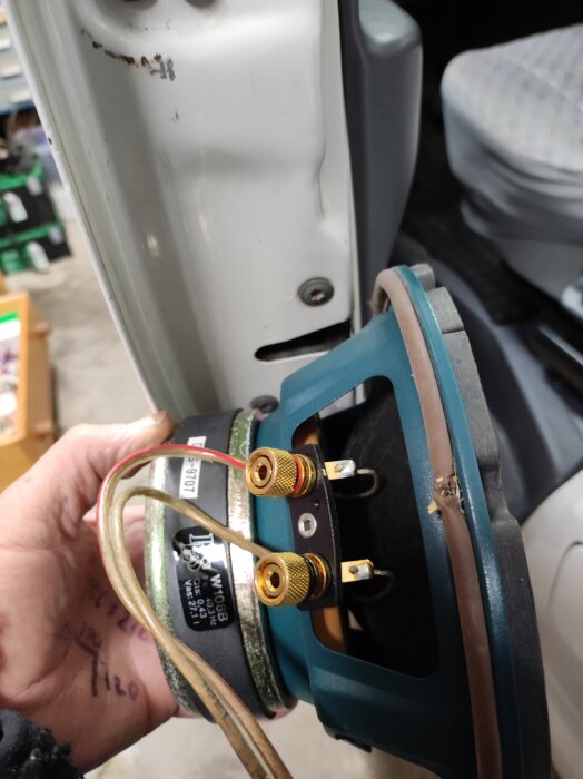 Hand håller högtalare som installationss i Toyota-bussdörr med synliga guldpläterade terminaler och kablar, monteringsutrymme problematiskt.