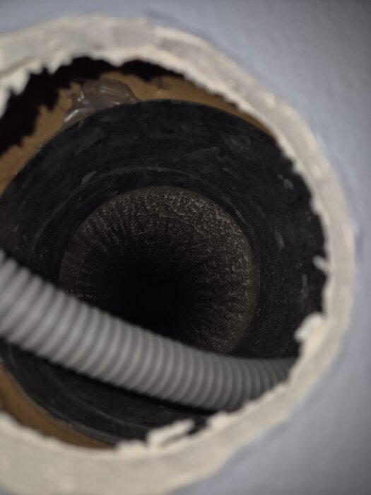Utsikt in i ett mörkt ventilationsrör med en grå, flexibel slang som löper genom hålet i väggen.