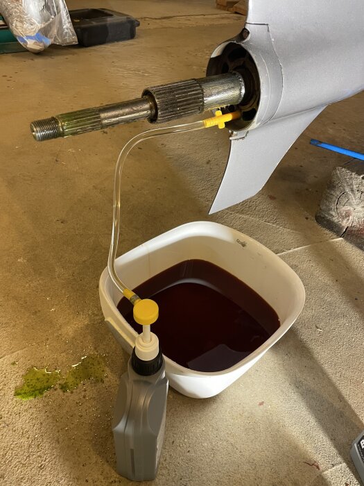 Bilden visar en båtdrivrem och drev där växelhusolja byts ut. En balja med röd vätska står under drevet, samt en liten flaska och en slang.