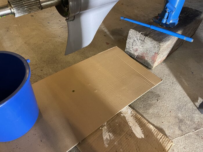 Träbåtsdrev med kartongskiva under för att upptäcka oljeläckage. Blå hink och träblock syns bredvid.