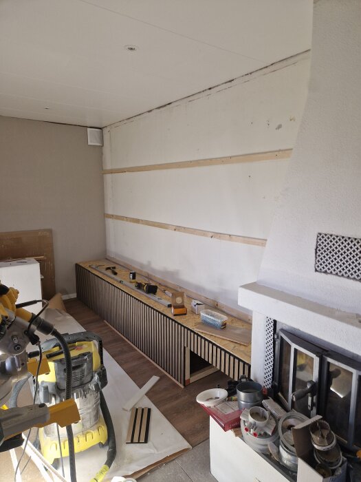 Platsbyggd tv-bänk under konstruktion med OSB och ekparkett, verktyg på toppen, nära en akustikvägg och öppen spis, med byggmaterial runtomkring.