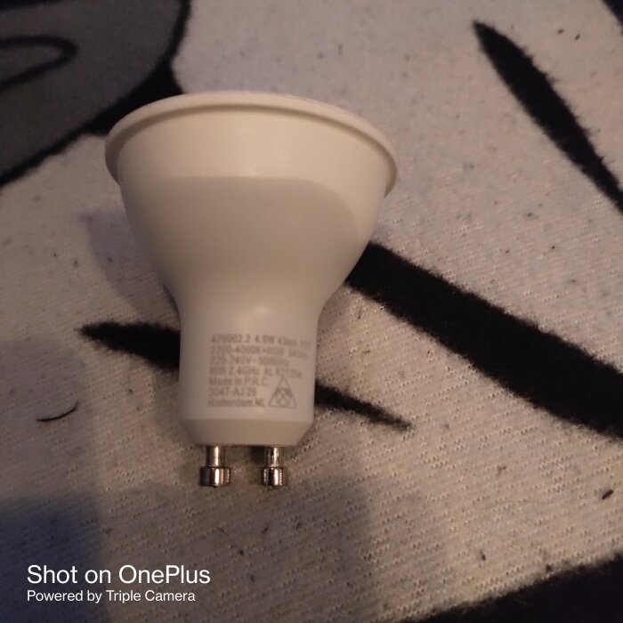 Vit GU10 LED-lampa med detaljerad text på basen, placerad på en svartvit tygbakgrund.