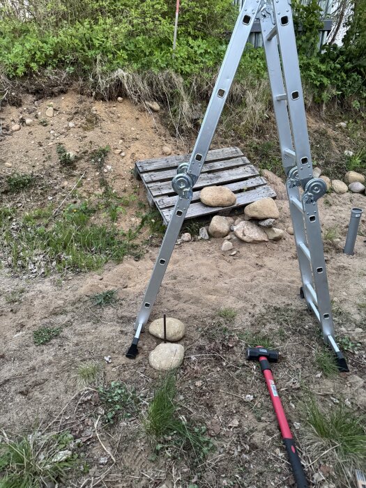Stege vid grävd grop, träpall och stenblock; armeringsjärn i marken och röd slägga på marken; gröna buskar i bakgrunden.
