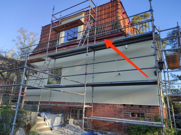 Byggnadsställningar runt ett hus med tegeltak under renovering. En röd pil pekar på avrinningsrännan under taket.
