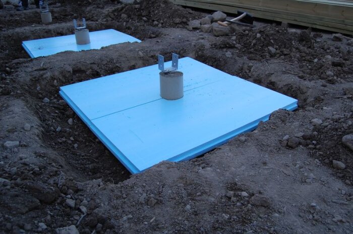 Isoleringsskivor av blått material i grävda hål i marken, med betongplintar som sticker upp. Används vid växthusbygge för att minska tjälskjutning.