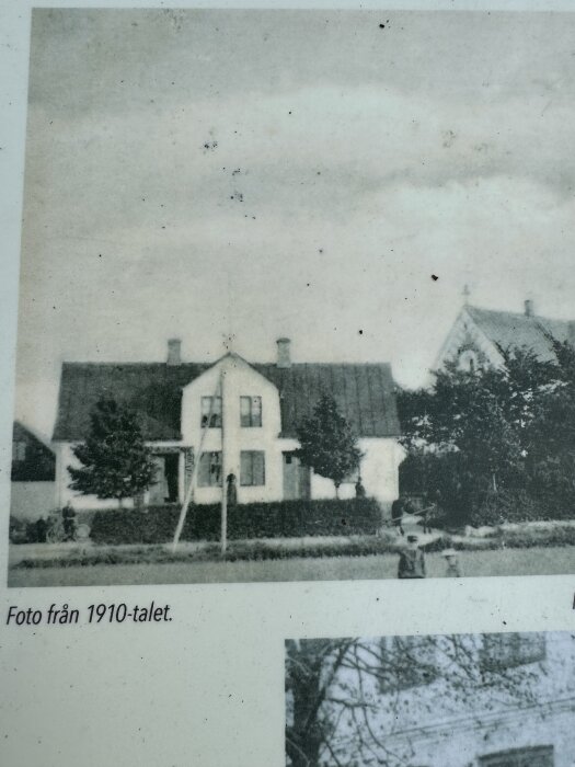 Ett gammalt hus med brant tak, omgiven av träd och staket, fotograferat runt år 1910.