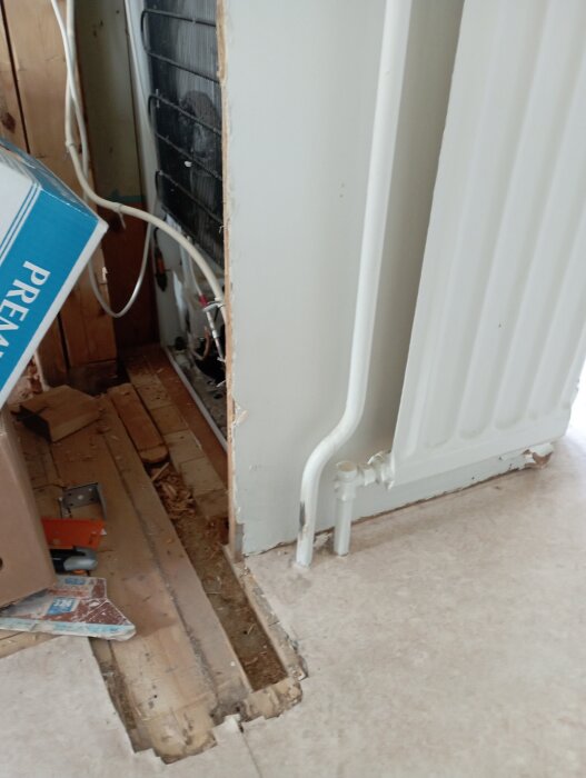 Radiator borttagen från vägg, exponerade rör och golv med öppningar, del av köksombyggnad.