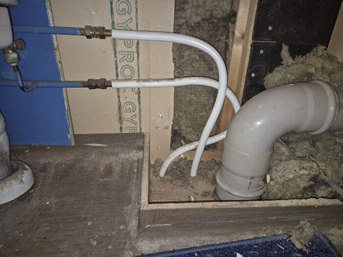 Vit avloppsrör i golvnivå med två vita vattenrör och isolering i ett badrumsrenoveringsprojekt.