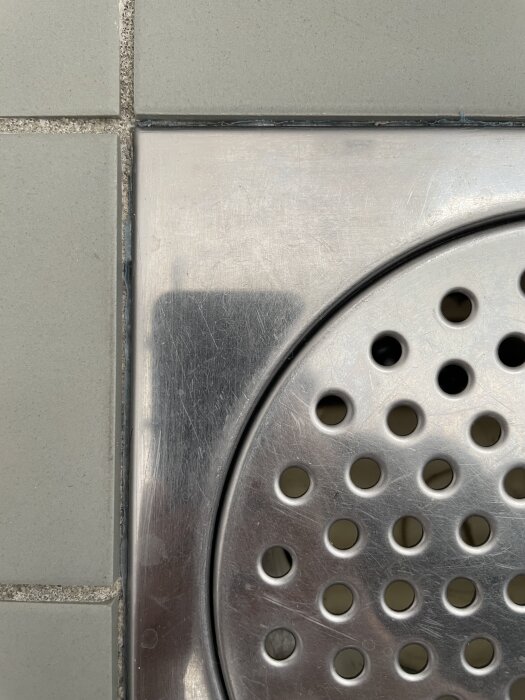 Grå klinker med blå plast som sticker ut vid golvbrunnen. Golvbrunn i metall med ihåliga klinker runtom.