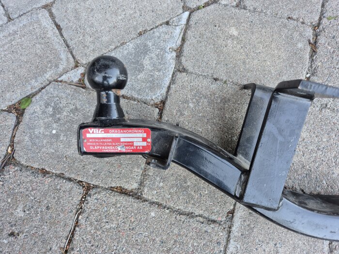 Dragkrok märkt "VBG draganordning" på stenbelagd mark, med en svart kula ovanpå.