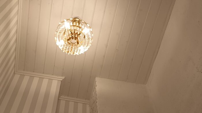 Tak med vitmålad träpanel och kristallampa i ett rum med randiga väggar.
