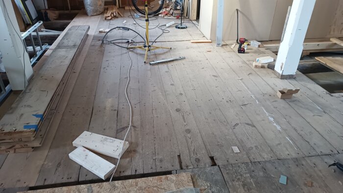 Trägolv under renovering med träplankor ligger på golvet, omgivna av byggmaterial och verktyg i ett pågående projekt.