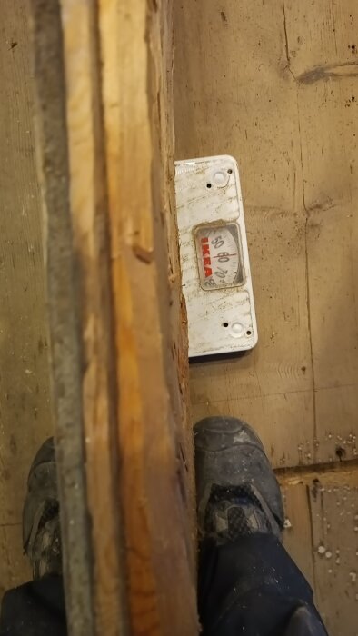 Närbild av en våg med en stor träplanka ovanpå, och ett par fötter i arbetsskor bredvid, på ett trägolv.