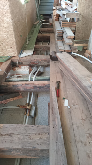 Installerat golv med synliga rör och staplade plankor på en byggarbetsplats. Verktyg och byggmaterial finns i närheten.