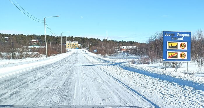 Snötäckt väg med hastighetsskyltar, vy mot samhälle, kall vinterdag. Vägenskylt på höger sida markerar gränsen till Finland.