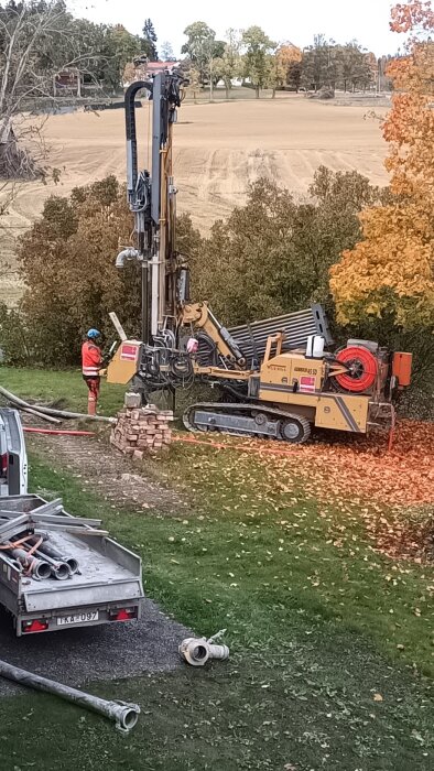 Borrigg för bergvärmeinstallation med en arbetare i orange skyddsutrustning står intill, omgiven av höstfärger och åkermark.