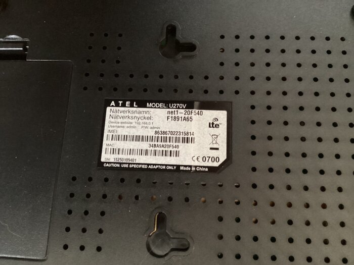 Närbild på en etikett på en Atel-modell U270V med nätverksnamn, nyckel, IMEI och MAC-adress visas på en routerns baksida.