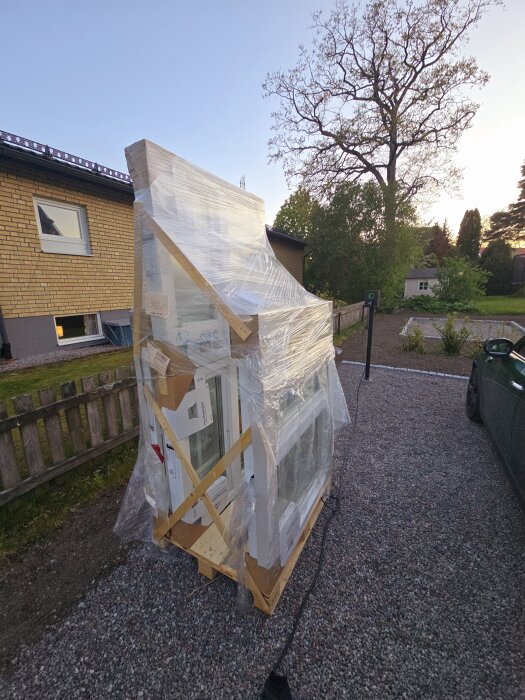 Fönster från Svenska Fönster inslagna i plast och emballage på en pall, placerade utomhus på en grusuppfart.
