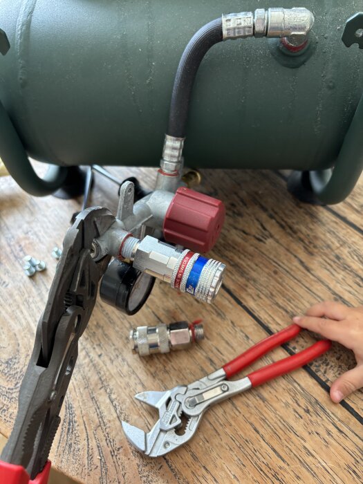 Barnsäkra anslutningar på en kompressor, med slang, kopplingar och verktyg som tång och skiftnyckel på ett träbord. Ett barns hand syns vid verktyget.