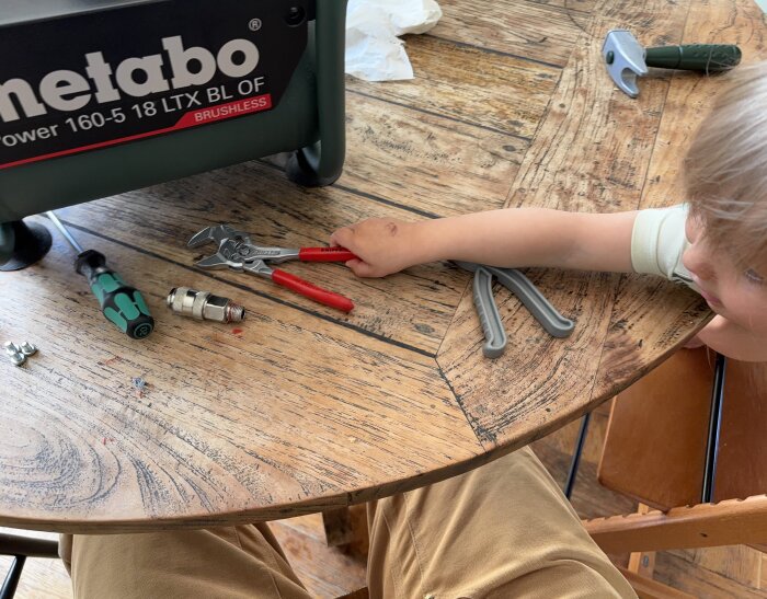 Barn vid bord med verktyg och en kompressor, bland annat en skruvmejsel och polygrip i närheten.