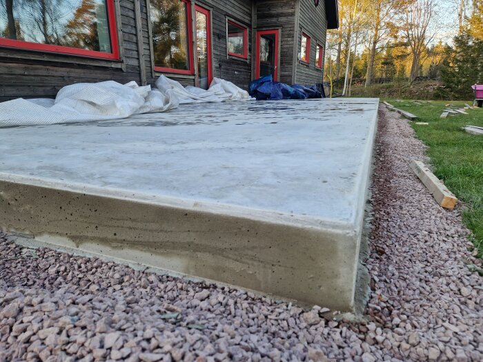 Betongplatta vid ett hus med borttagna formar och avrundade kanter, omgiven av grus och gräs.
