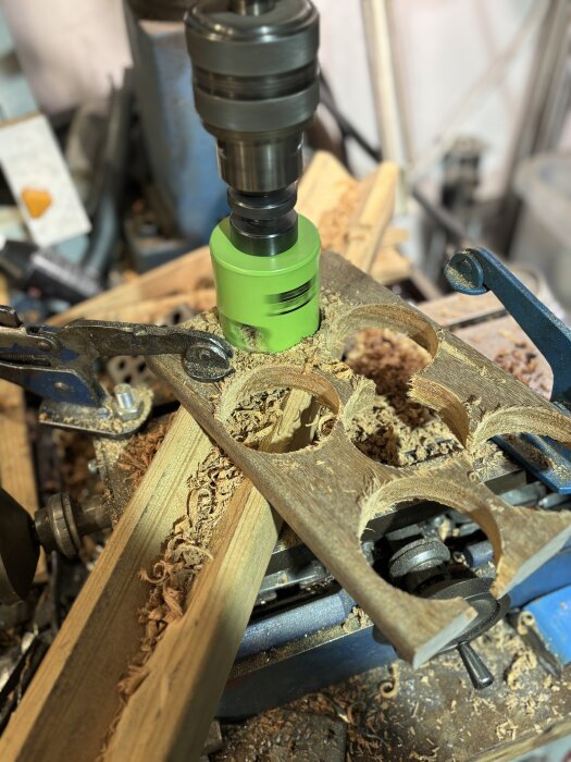 Borrmaskin med hålsåg skapar runda hål i träplanka, omgiven av träspån och verktyg i en verkstadsmiljö.