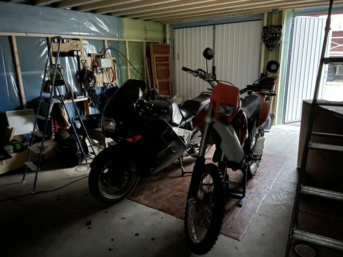 Två motorcyklar i ett garage, omgivna av verktyg och en stege, med garageporten delvis öppen i bakgrunden.