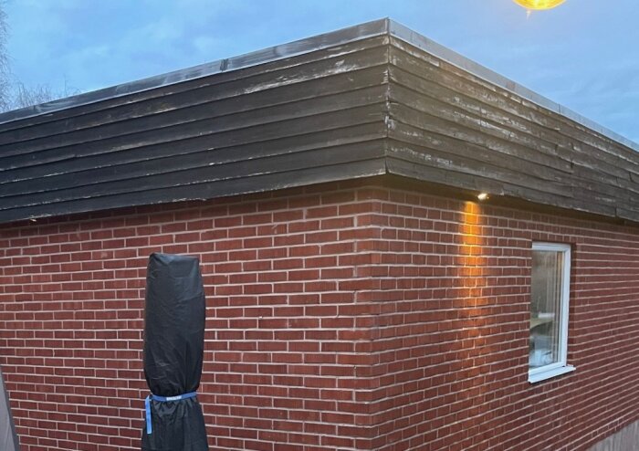 Rött tegelhus med svart panel upptill, belysning vid ett av hörnen. Plåtarna vid takets ytterhörn ska bytas.