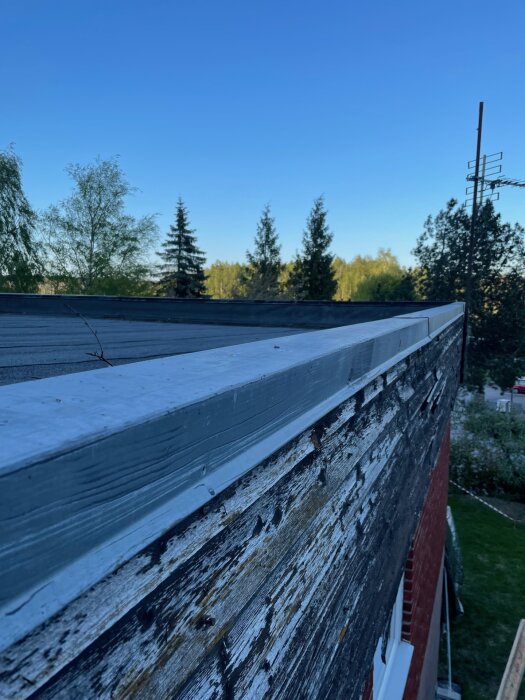 Ytterhörn på ett hus med sliten panel och plåtinfattning mellan vägg och tak, omgiven av träd och klarblå himmel.