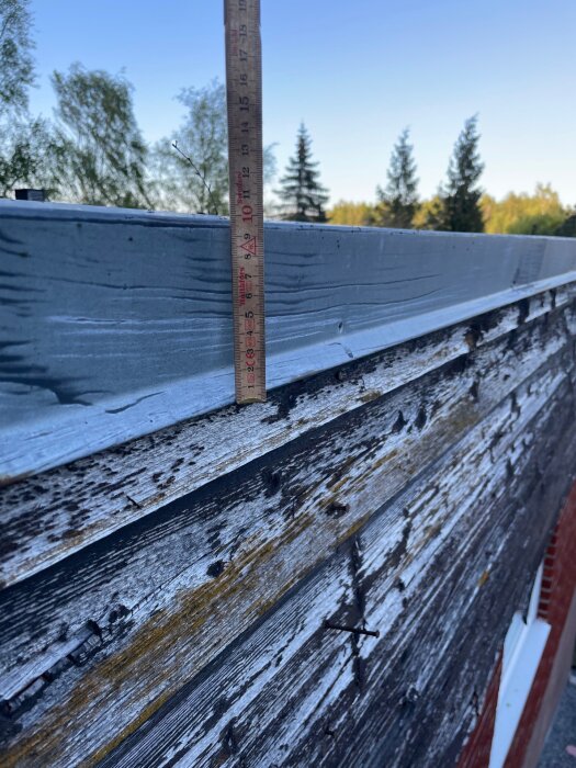 Sliten träpanel på hus med lodrätt mätsticka mot takplåt, omgiven av träd och blå himmel i bakgrunden.