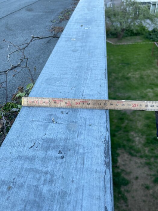 Måttstock på plåtbeslag vid husets hörn, del av takets kant, med gräs och träd i bakgrunden.