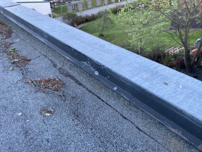 En kant av tak med grå plåt, täckt av skräp och grenar, mot bakgrund av en grön trädgård.
