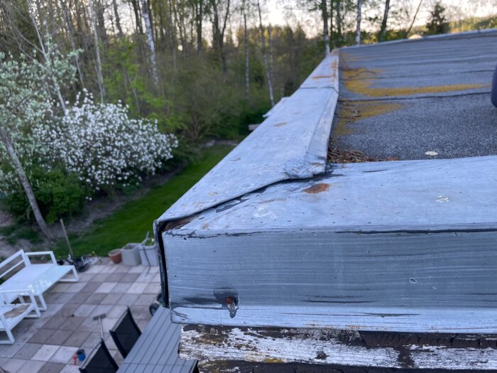 Plåtinklädda ytterhörn på takkant med antydan till rost och mossa, trädgårdsmiljö i bakgrunden.