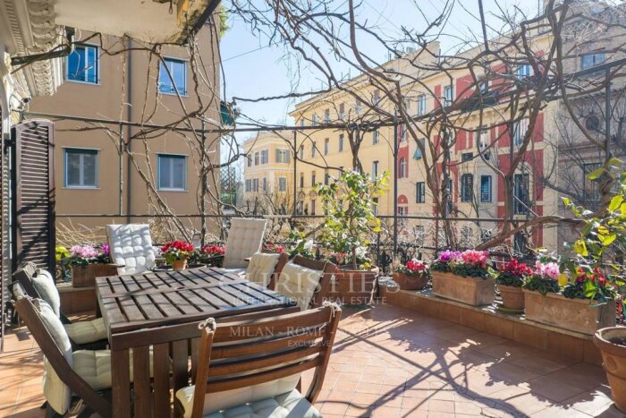 Italiensk terrass med trämöbler, växter i krukor och utsikt över pastellfärgade byggnader med dekorationer i rött och valv.