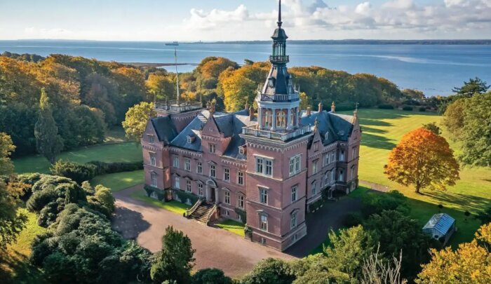 Storslaget gods vid vatten omgivet av höstklädda träd och stor gräsmatta. Historisk byggnad med torn och utsikt över vattnet.
