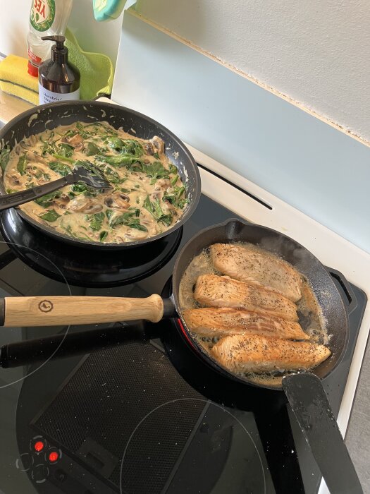 Stekt lax i stekpanna och ostsås med lök, svamp och spenat i en annan panna på spis.