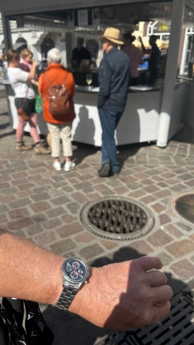 Man med armbandsur i förgrunden, folk vid vinstånd på torg i Trier i bakgrunden.