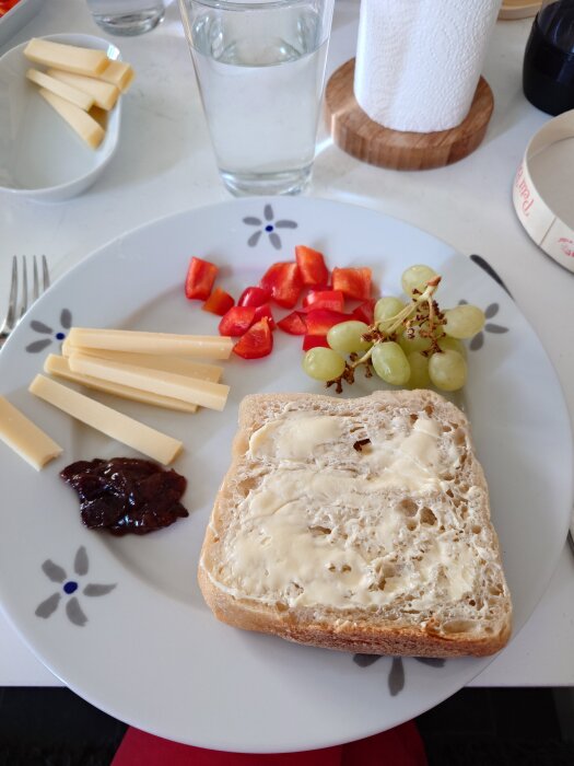 Tallrik med bröd med smör, oststavar, hackad paprika, vindruvor och en klick marmelad, samt ett glas vatten.