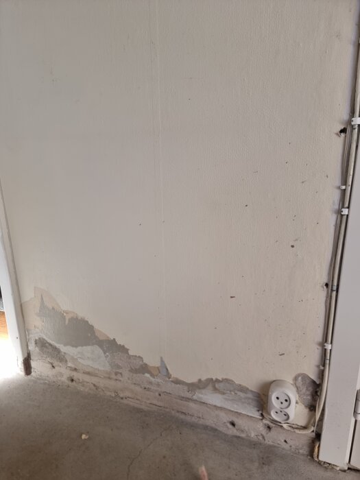 Vägg med delvis borttagen tapet och synlig puts i en källare. Elektriskt uttag och skadad nedre del av väggen.