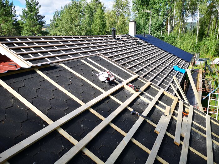 Tak under renovering med träreglar på asfaltschingel, verktyg utspridda. Skorsten och natur runt omkring.