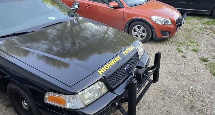 Svart "Highway Patrol"-bil med push bar monterad, stående bredvid en röd bil på en grusplan.