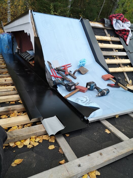 Takrenovering med verktyg och material på plats, inklusive takpapp och plåt. Löv och trästommar syns i bakgrunden.