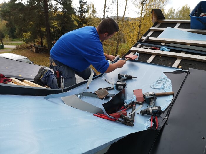 Person arbetar på tak med verktyg och material, förbereder takgenomföringar och skorsten. Takarbete omgiven av höstlandskap i bakgrunden.